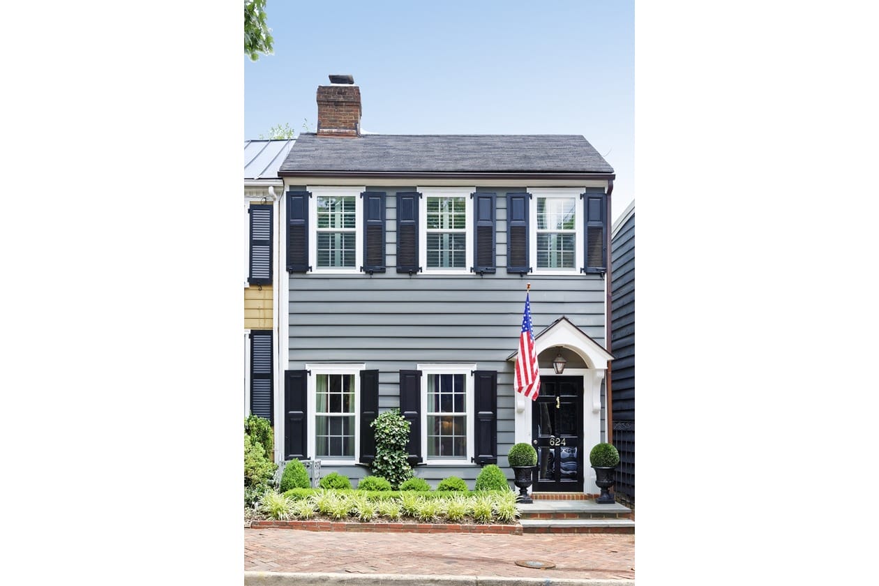 Two-story gray-blue townhouse with black shutters, white trim, and an American flag at the front door amid neatly trimmed shrubs on a brick sidewalk