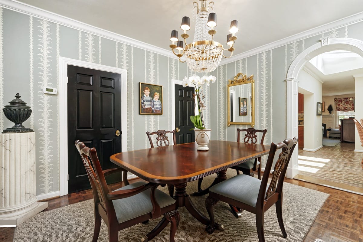 Formal dining room with an oval mahogany table, carved chairs, and a crystal chandelier. An elegant, patterned wallpapered wall behind.