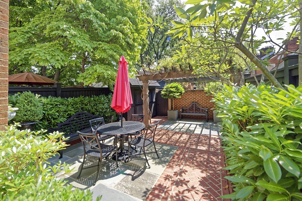 Outdoor brick patio with a round metal table, six chairs, and a red umbrella among green hedges and trees.