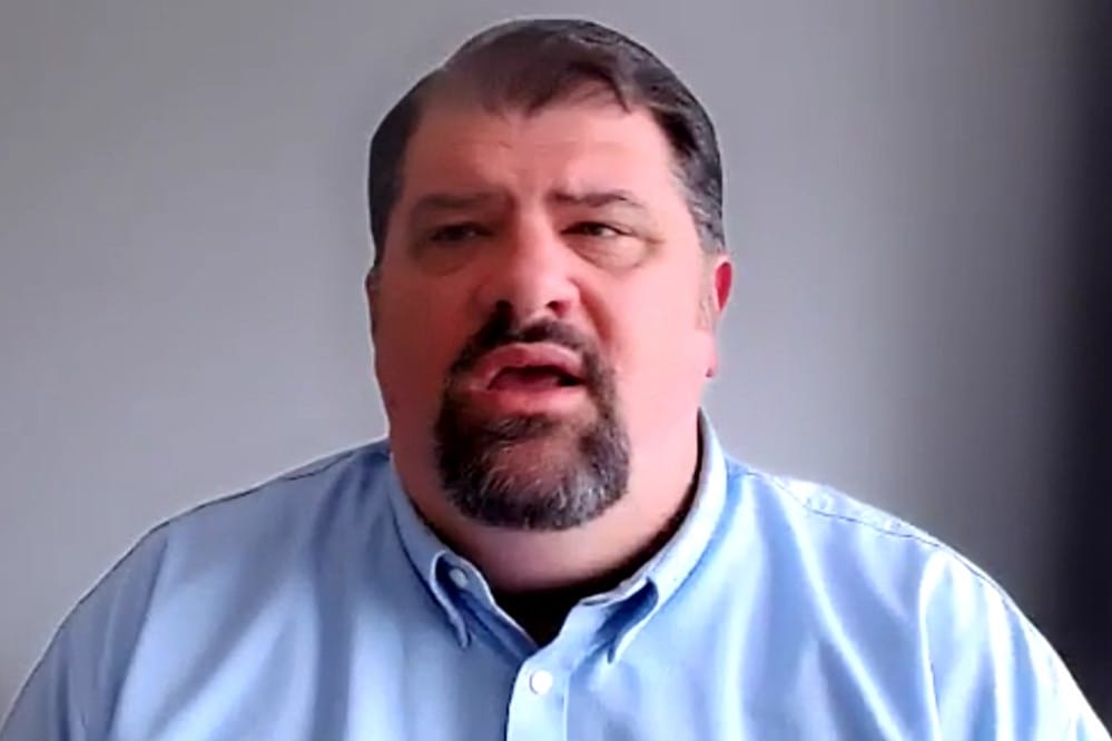 Man with short dark hair and goatee wearing a light blue button-up shirt speaking during a video call.