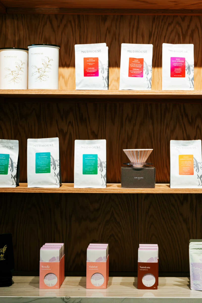 Shelf display featuring white Mudhouse coffee bags with colorful labels on wooden shelves, plus a dark box with a cone-shaped accessory in the middle row and orange boxes on the bottom row.