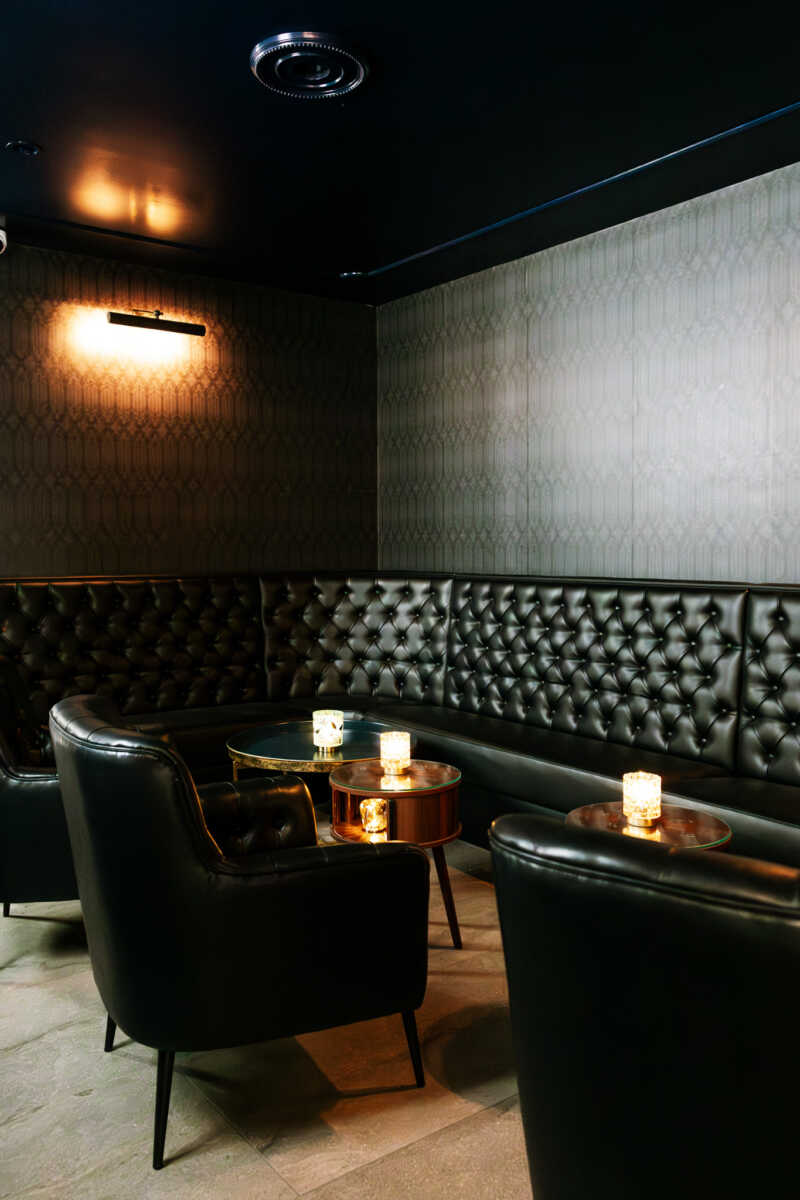 Cozy lounge area with black tufted leather seating and small round tables lit by candlelight in a dim room.