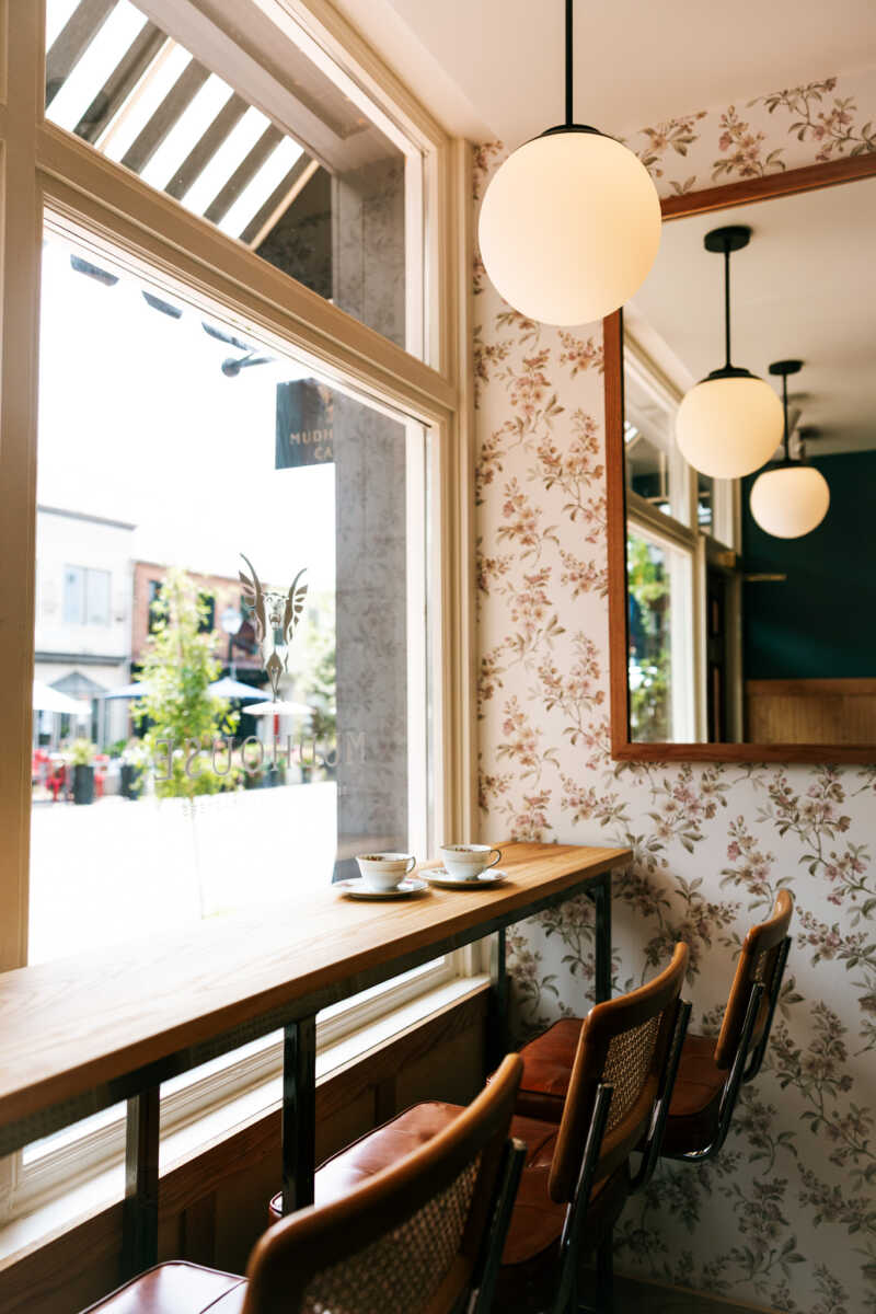 Cafe interior by a large window with two tea cups on a wooden counter, floral wallpaper, and round pendant lights.