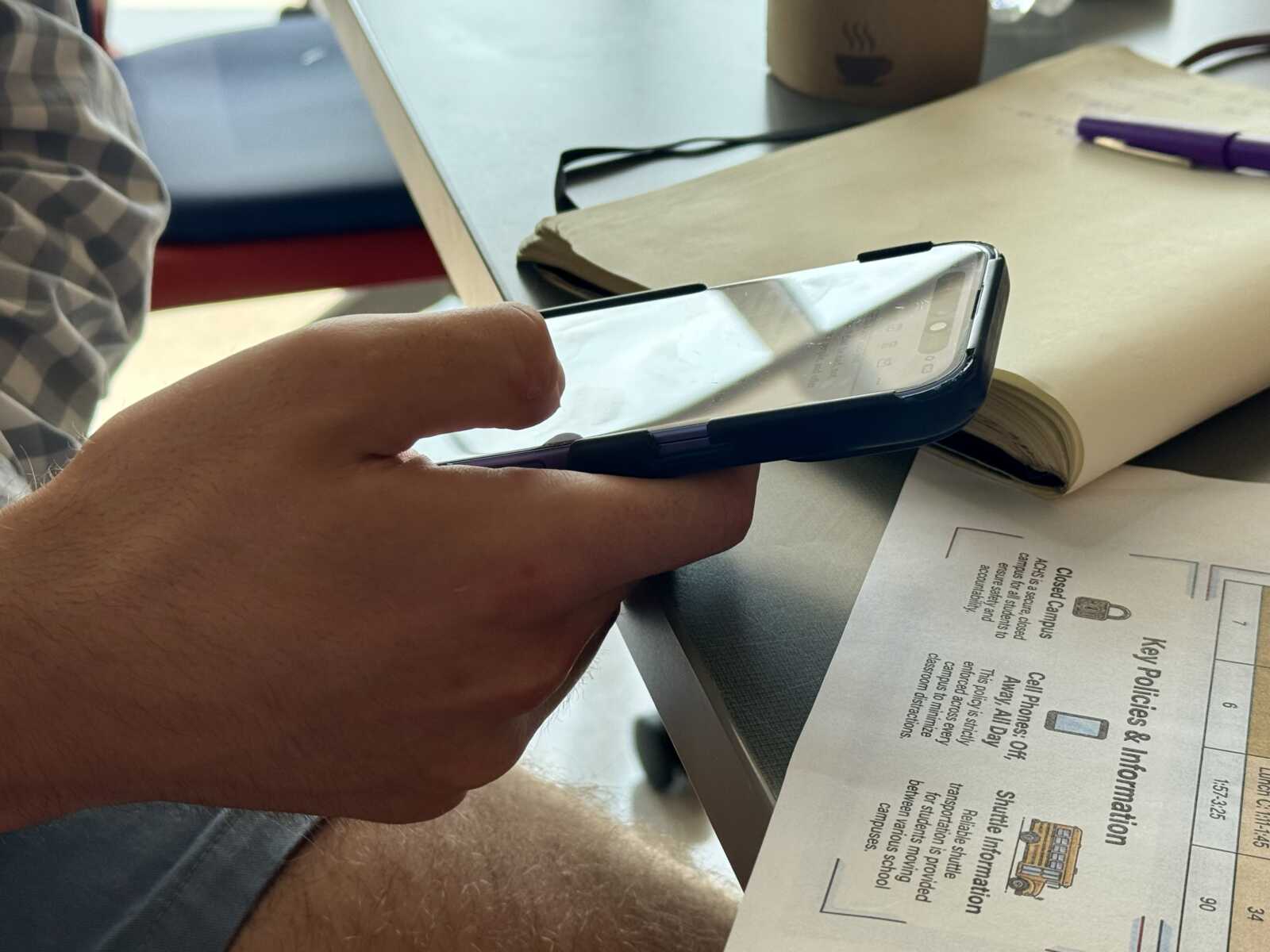 Person holding a smartphone in landscape orientation over a desk with papers and a notebook nearby.