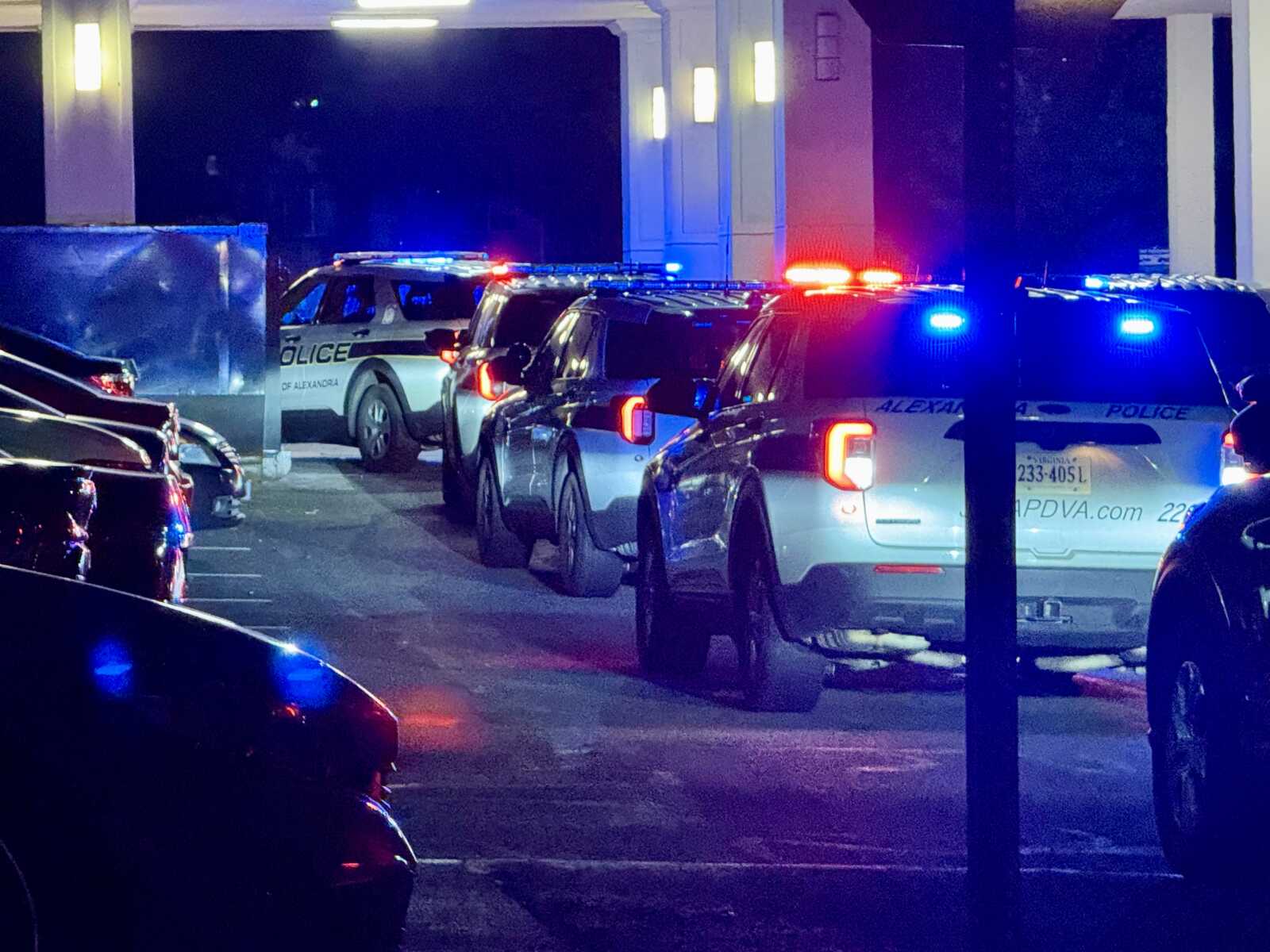 Line of police SUVs with flashing red and blue lights at night in a parking area, suggesting an active police scene under a porte-cochère.