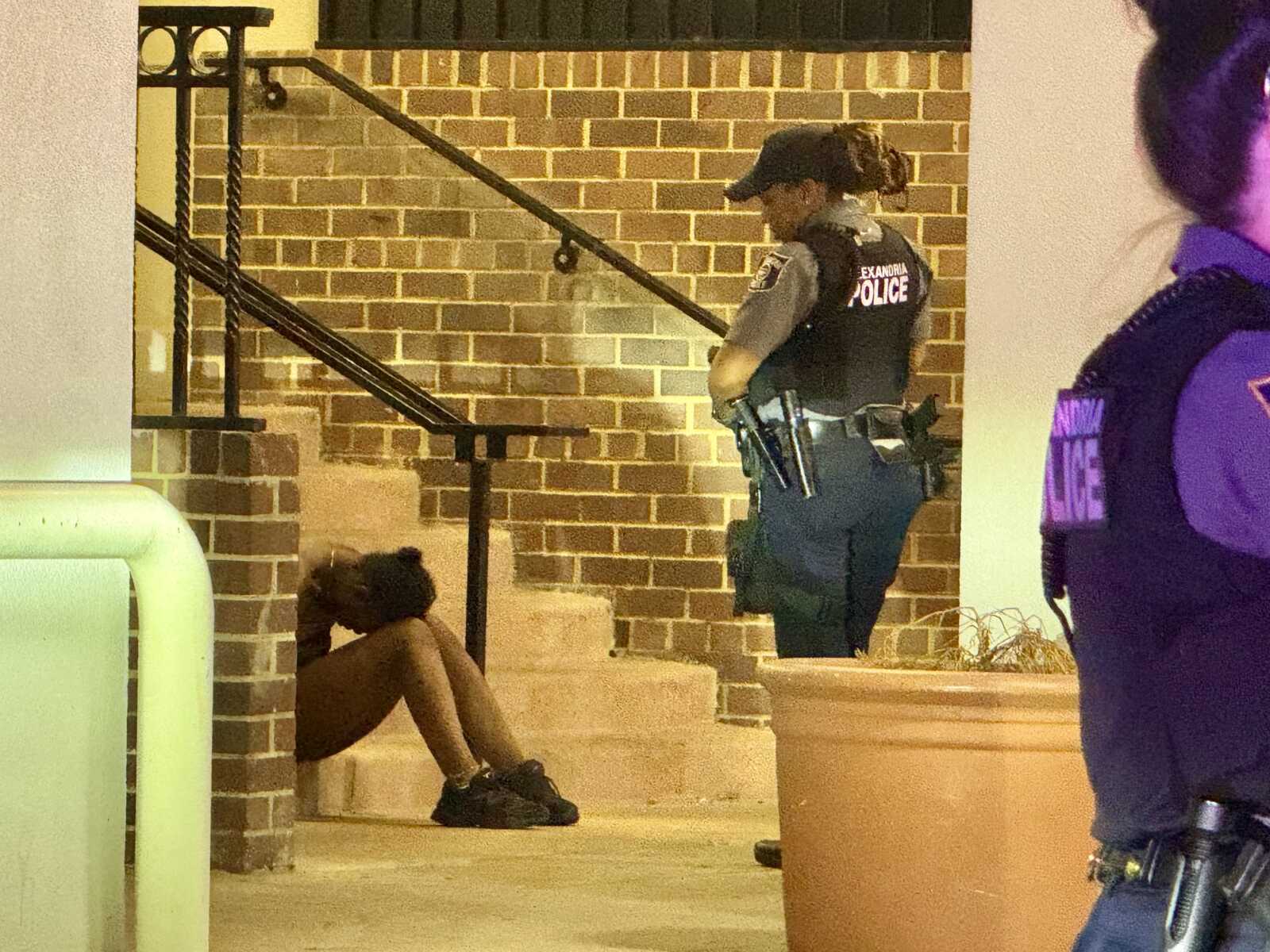 A police officer in a tactical vest stands by a seated person on brick steps at night, with another officer partially visible on the right.