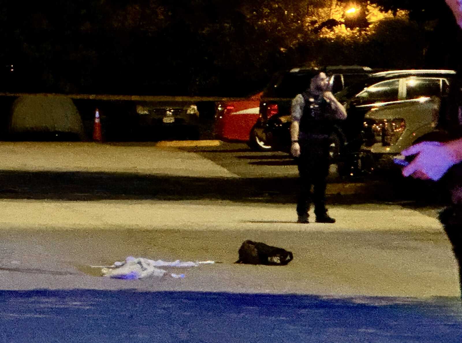 Nighttime street scene with a police officer standing in the foreground and vehicles in the background near a cordoned area and traffic cones.