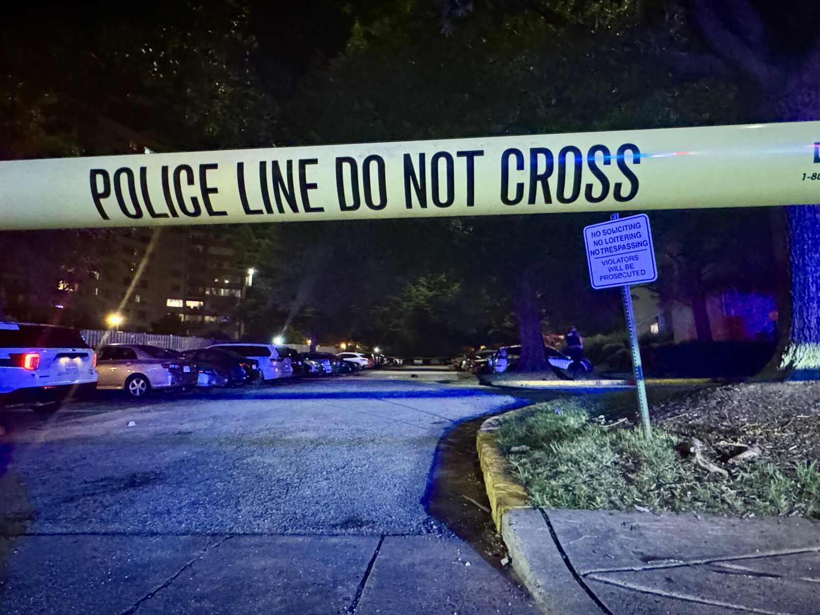 Nighttime scene with police tape reading 'POLICE LINE DO NOT CROSS' stretched across a street and rows of parked cars behind it.