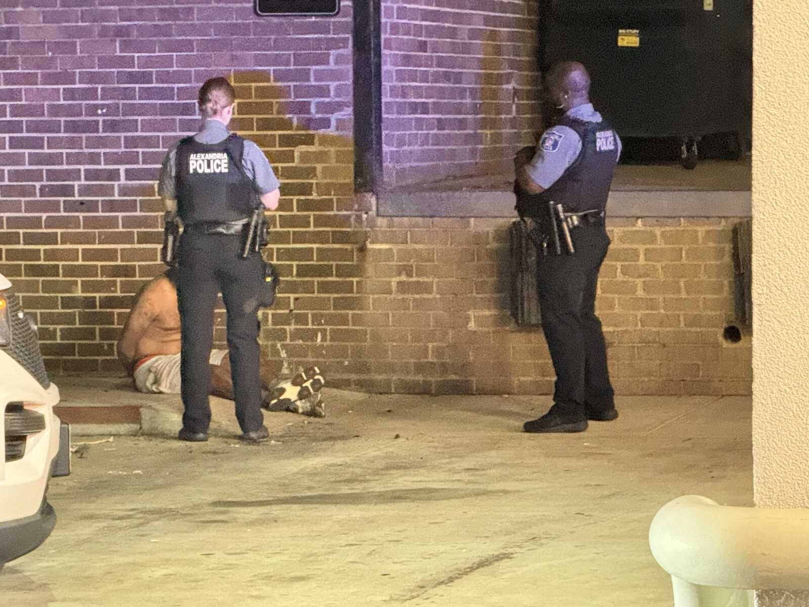 Two police officers stand guard near a shirtless man sitting on the curb against a brick wall at nightfall.