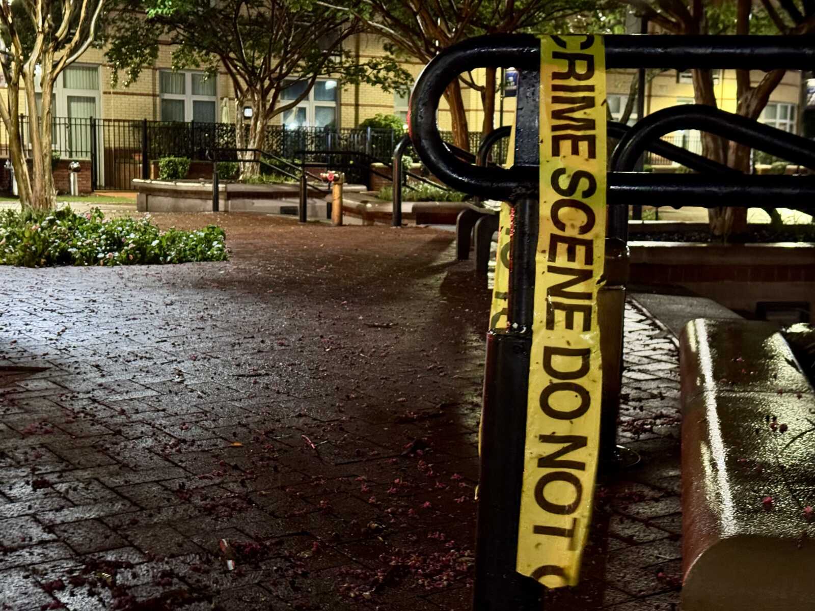 Yellow crime scene tape wrapped around a black railing in a wet courtyard at night, cordoning off the area.