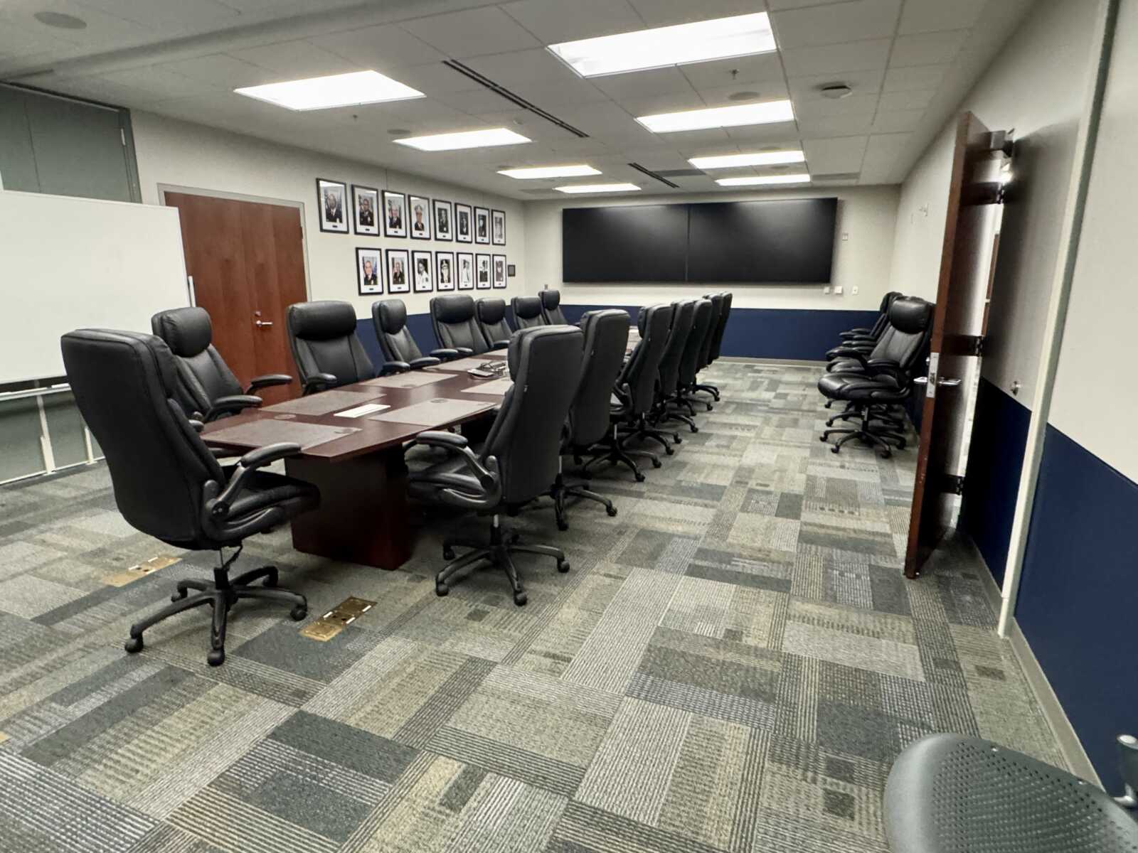 A modern boardroom with a long wooden table and black leather executive chairs around it; portraits line the wall and a large dual-screen display at the front.