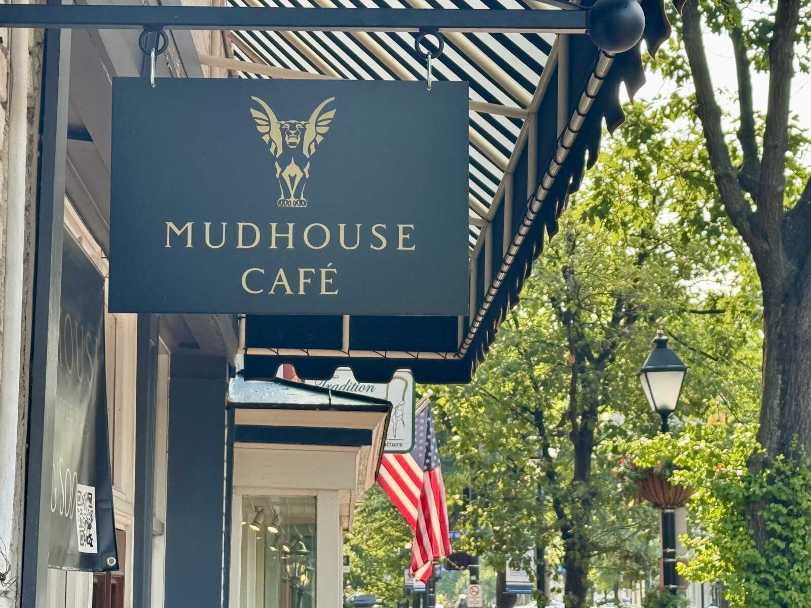 Blue storefront sign reading 'MUDHOUSE CAFÉ' with a winged creature logo hanging above a sidewalk.