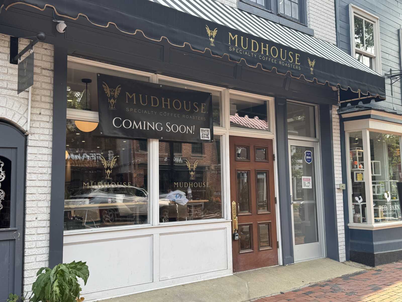 Mudhouse storefront with black awning and banners announcing 'Coming Soon!' above large windows and a wooden door.