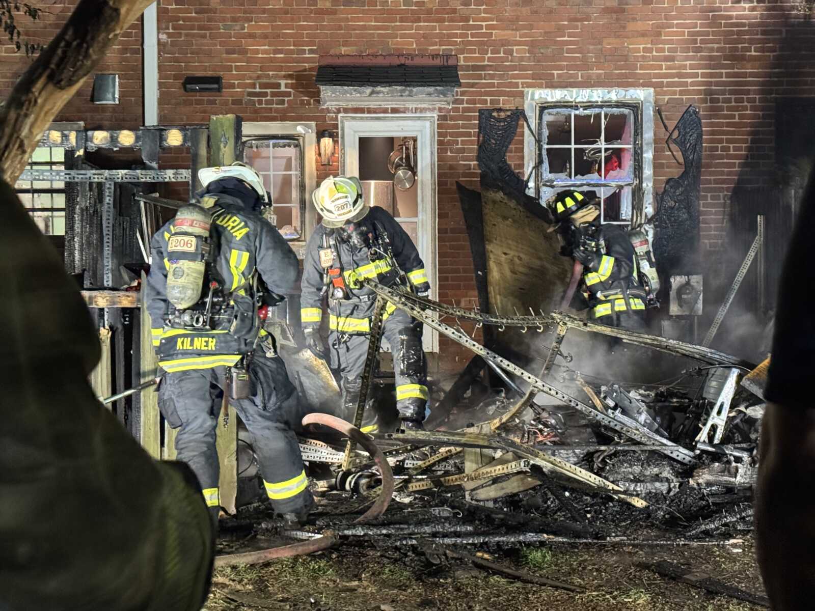 Firefighters in full turnout gear working amid a burned brick house with charred debris and tangled hoses, smoke rising.