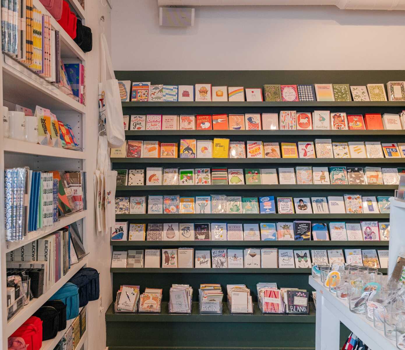 Interior of a shop displaying rows of colorful greeting cards on green shelves along the back wall, with shelves of notebooks on the left.