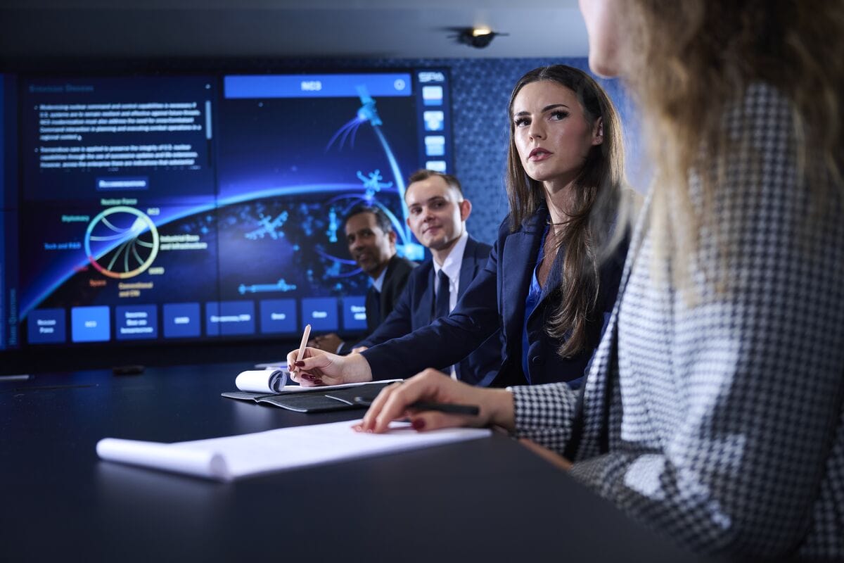 Business meeting with a woman in the foreground taking notes while colleagues review data on a large monitor in a high-tech boardroom.