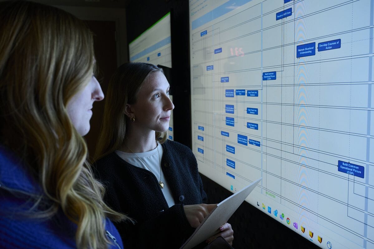 Two professionals study a large wall display showing a blue timelines chart, one holding a folder.