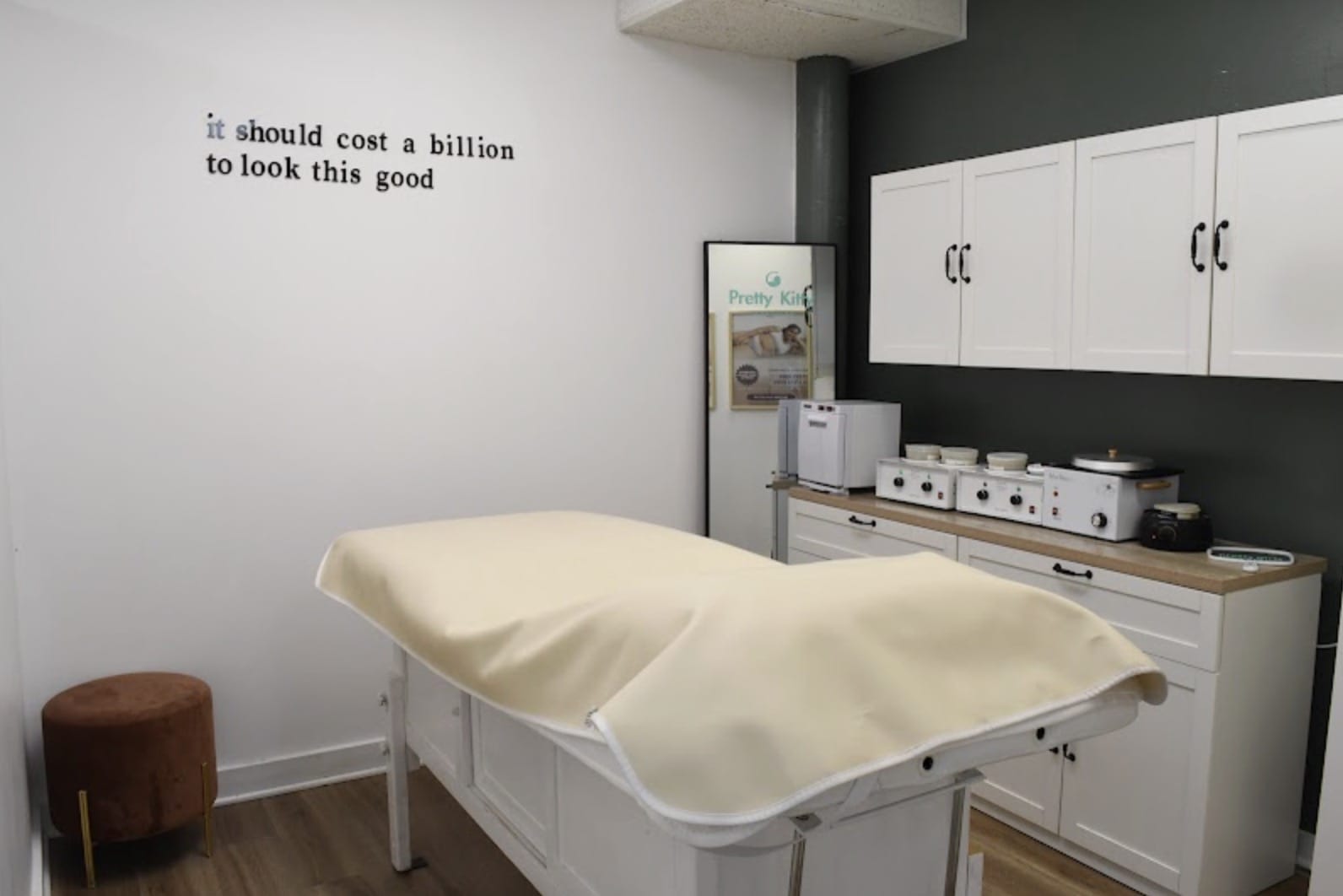 Treatment room with a padded examination table covered in a cream sheet, white cabinets, and small appliances on the counter.