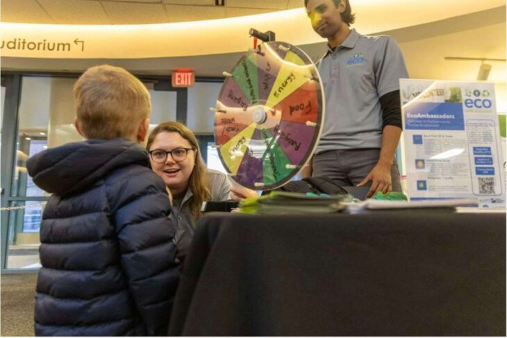 Woman with glasses at an Eco event smiles at a child while a colorful prize wheel spins at a booth in a campus lobby area.
