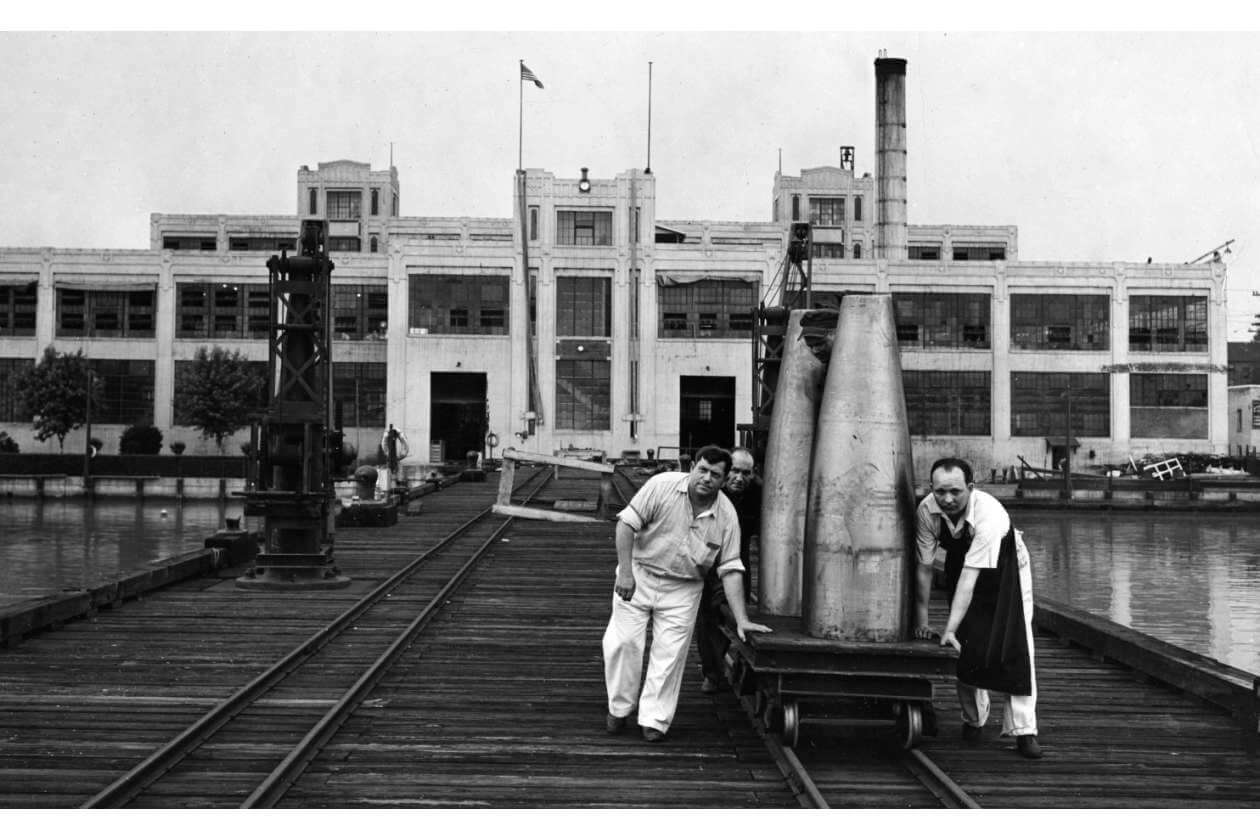 Black-and-white photo of three men wheeling a platform with a large cone-shaped metal object along railroad tracks at a waterfront factory yard.
