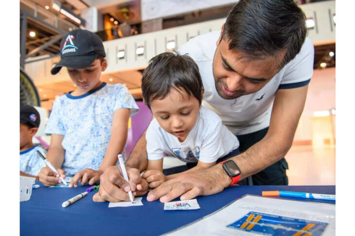 Man helping a child draw at a blue-table activity with markers in a bright indoor space while another boy watches nearby