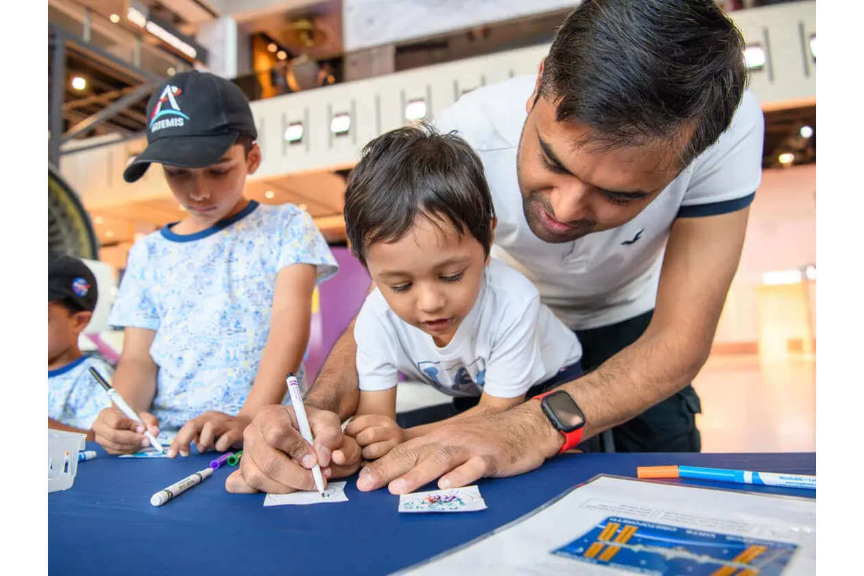 Man helping a child draw at a blue-table activity with markers in a bright indoor space while another boy watches nearby