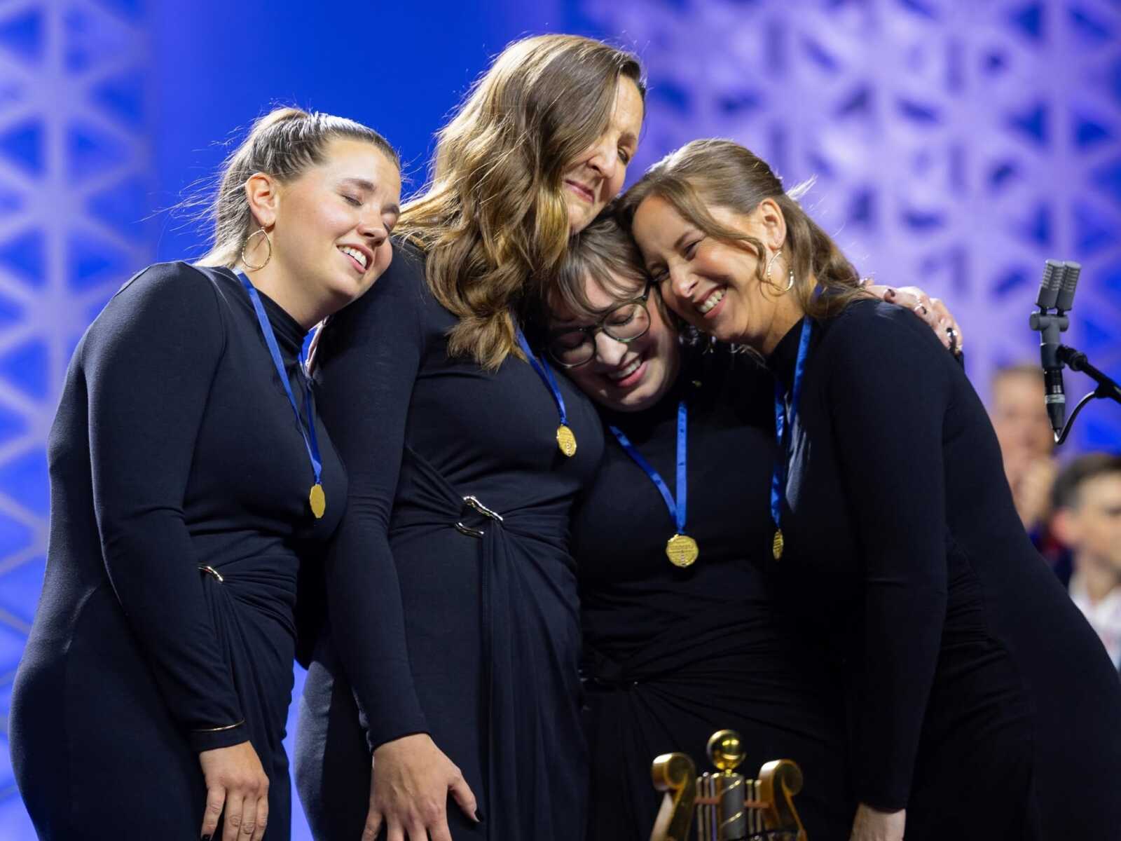 Four women in black dresses hug on a blue-lit stage, wearing gold medals around their necks with a trophy in front, celebrating.