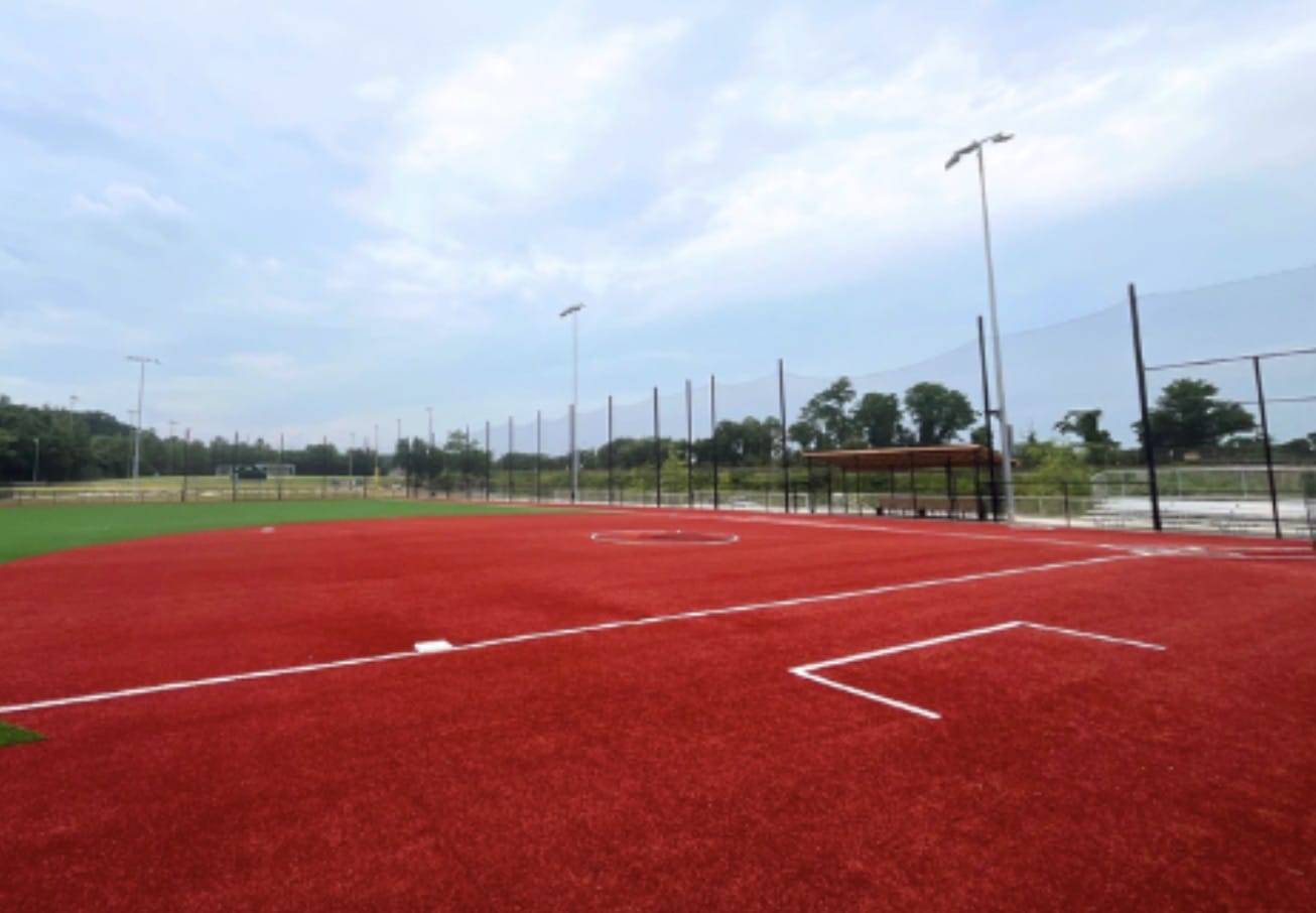 Red artificial-turf baseball infield with white baselines, fence, and a small dugout under a cloudy sky