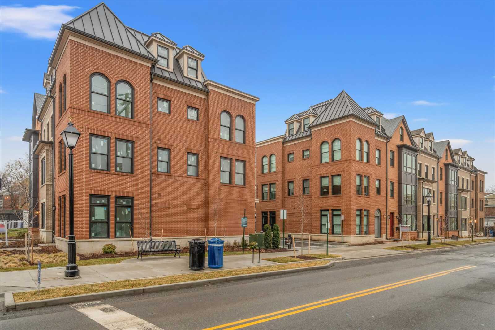Red brick townhouse-style apartment buildings line a street under a clear blue sky, with arched windows, dormer roofs, and black street lamps.