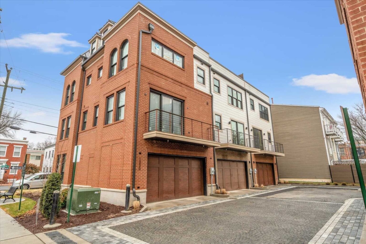 Three-story brick townhouse with brown garage doors and metal balcony railings on a sunny street.