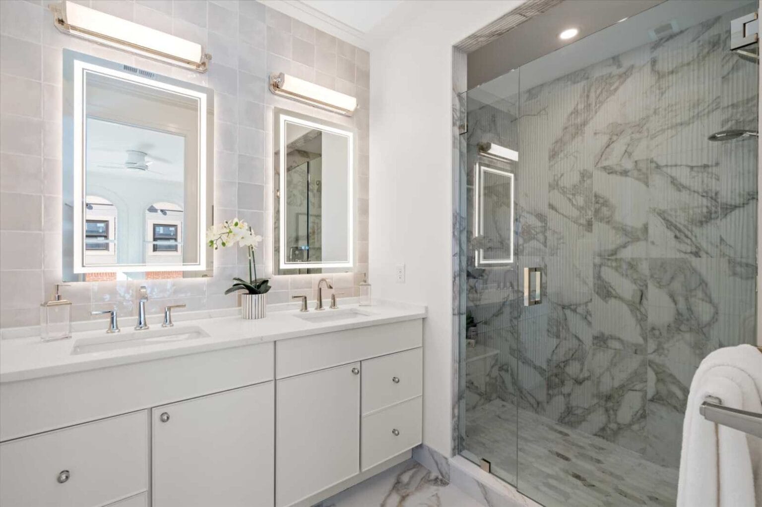 White double vanity with two illuminated backlit mirrors and chrome fixtures, marble-tiled shower in a glass enclosure.
