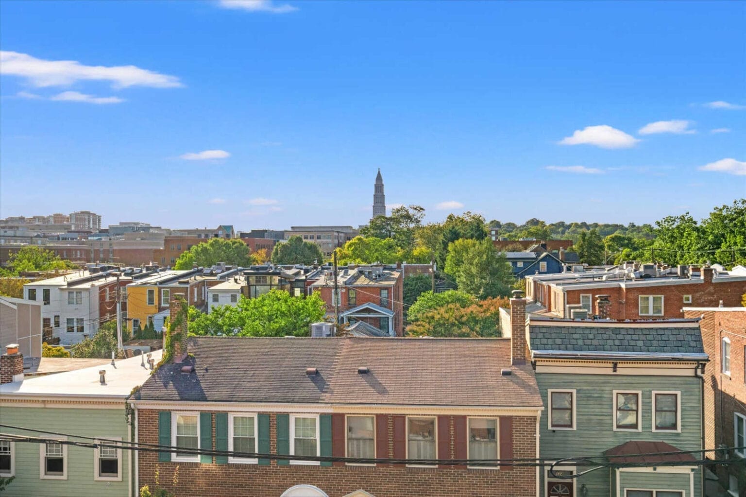 A row of colorful townhouses with green shutters in the foreground and a skyline with a tall historic bell tower in the distance under a bright blue sky.