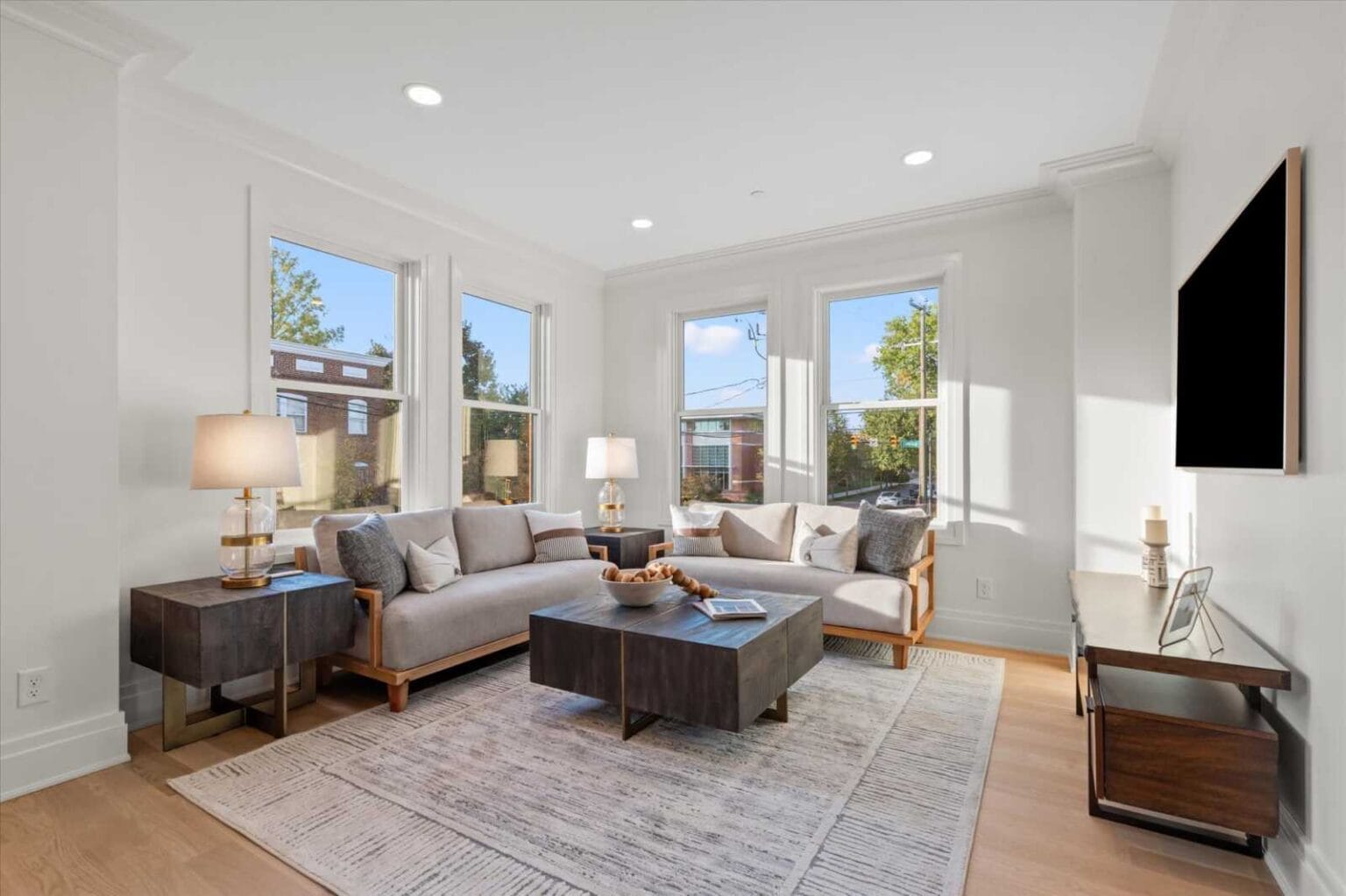 Bright modern living room with a light gray sectional, dark wood coffee tables, and large windows letting in sunlight, plus a wall-mounted TV on the right.