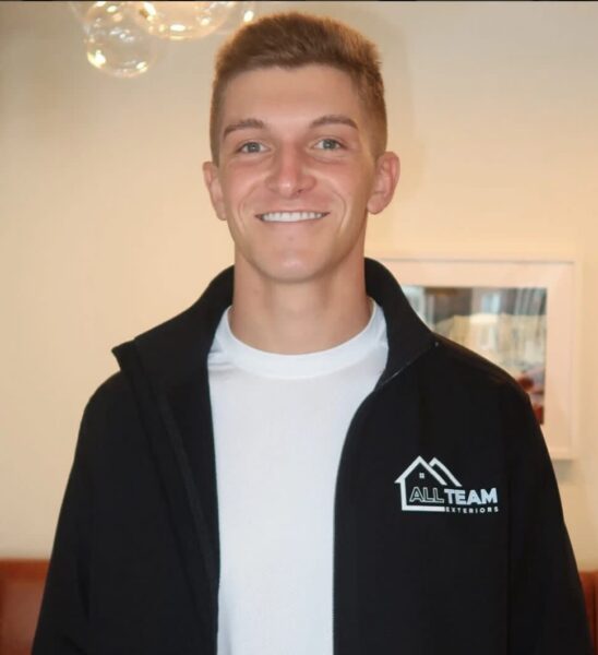 Smiling man in a black jacket with the All Team Exteriors logo, indoors in a casual setting.