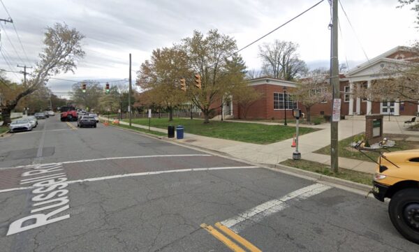 Street intersection near a brick campus building; traffic lights overhead, cars on the street, and a yellow school bus at the right edge.