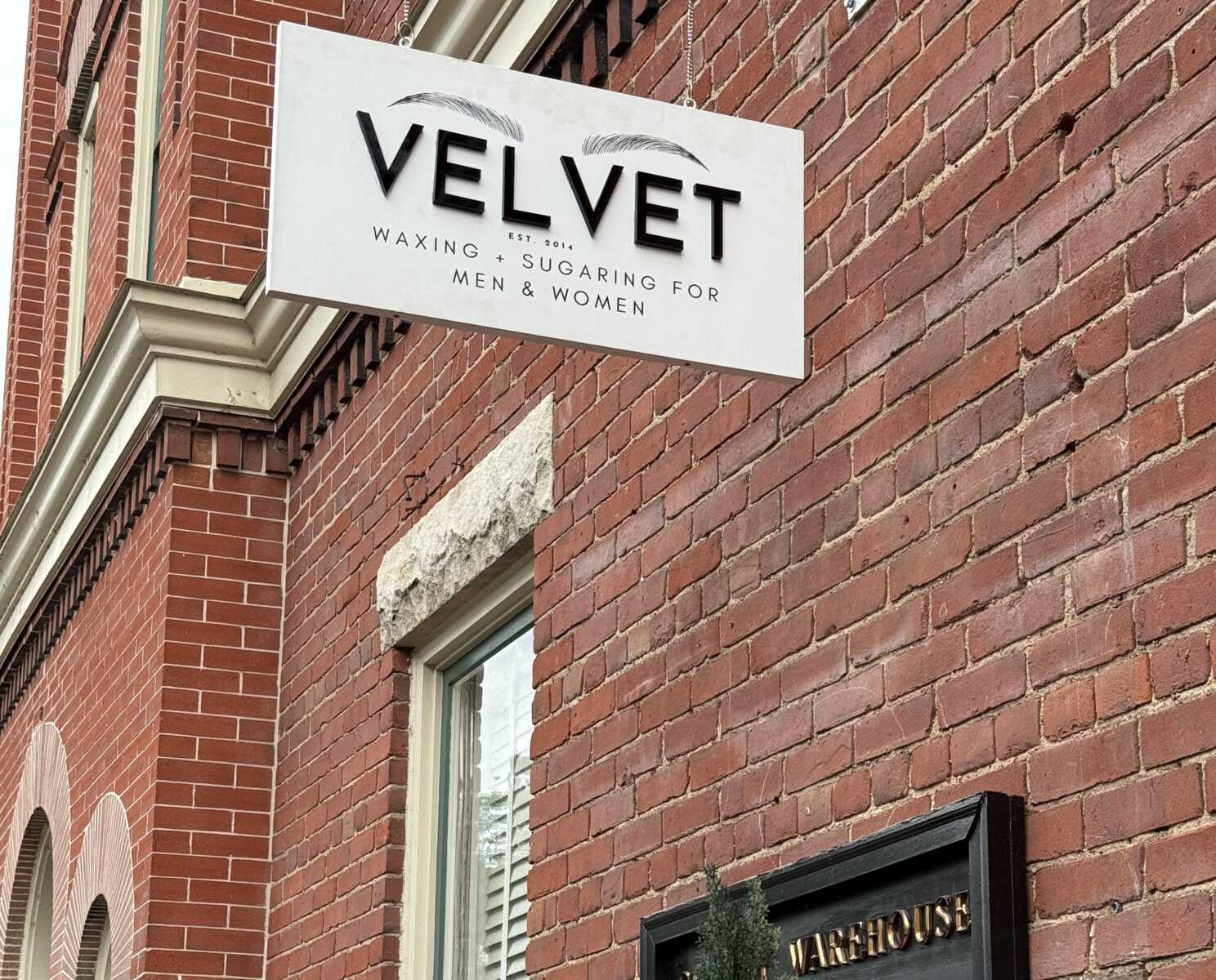 White rectangular sign reading 'VELVET' advertising waxing and sugaring for men and women on a red brick storefront edge.