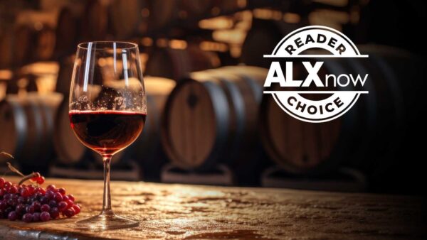 Wine glass of red wine on a wooden bar with grapes nearby and wine barrels in the blurred background, featuring a 'Reader ALX Now Choice' logo overlay.
