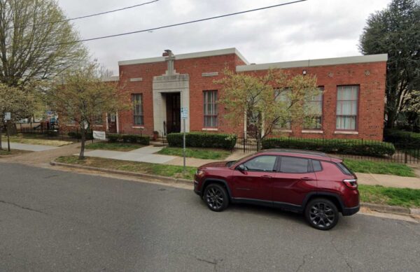 Brick, single-story building with a stone entryway and cross, front lawn with small trees, and a maroon SUV parked along the curb on a residential street.