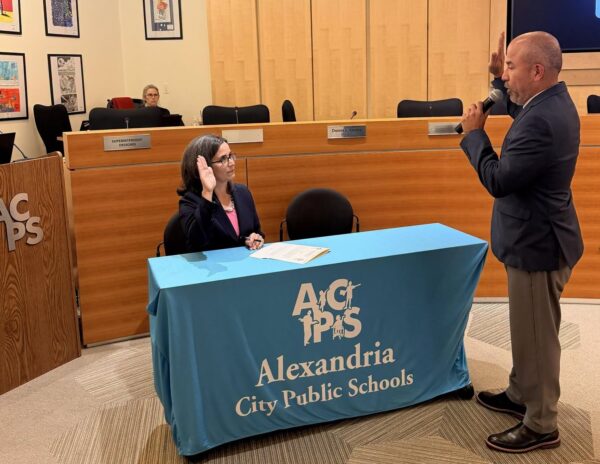 School board meeting: a woman raises her right hand to take an oath at a table with a blue cover that reads Alexandria City Public Schools, as a man speaks into a microphone beside her.