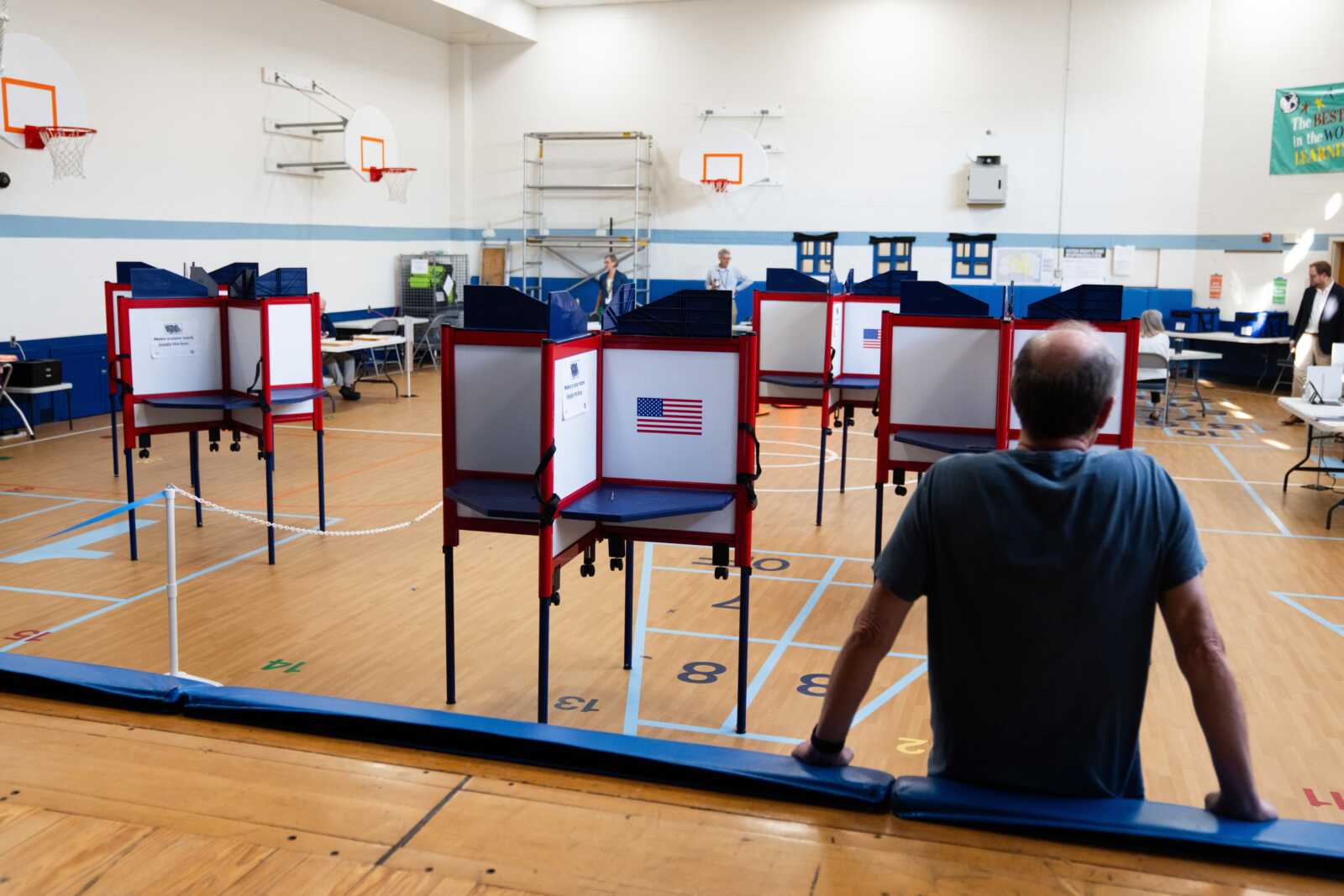 A man sits with his back to the camera in a gym turned polling station, red voting booths set up for voting.