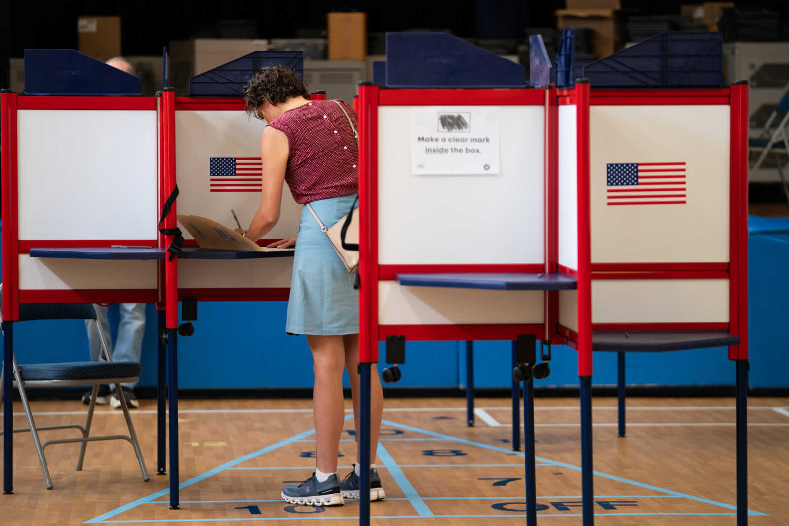 Person voting at a polling station inside a gym, using a red-and-white voting booth with American flags on the panels.