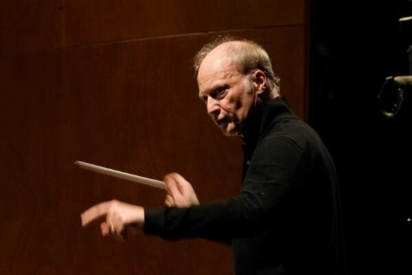 Conductor in black shirt using a baton to cue an orchestra on a dim stage.