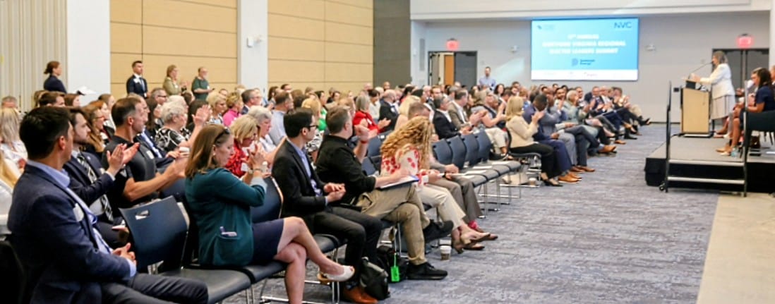 Audience applauding while a speaker presents on a stage in a conference room with a projection screen in the background.