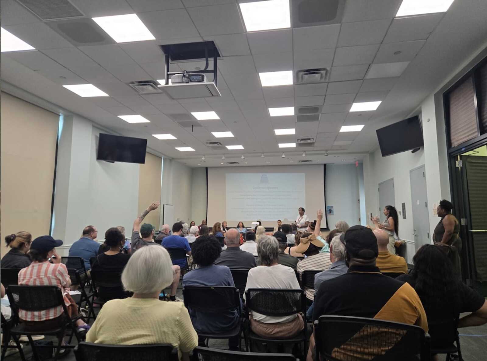 Audience seated in a conference room facing a panel at the front with a projector screen displaying slides.