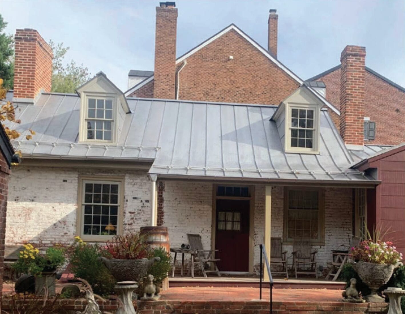 Front view of a brick house with a gray metal standing-seam roof, two dormer windows, and a covered porch with chairs and plants.