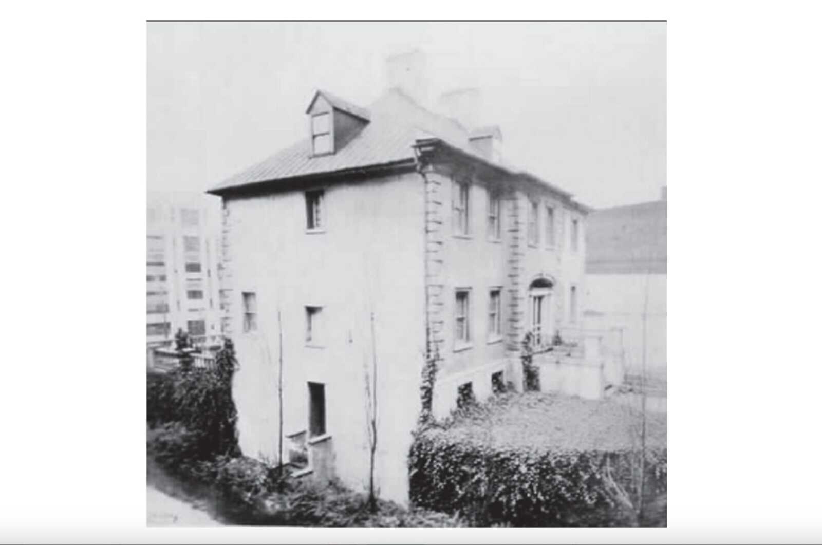 Three-story house with stone corner quoins, dormer window, and ivy-covered front yard on a sloped street (historical black-and-white photo)