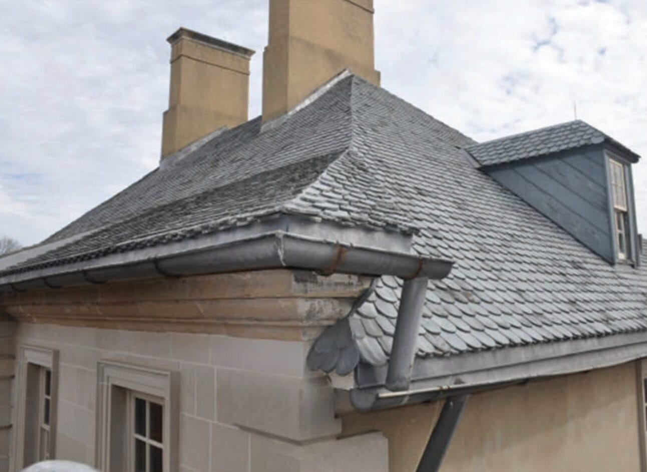 Slate-tiled roof with dormer and metal gutters on a stone building, chimneys in the background.