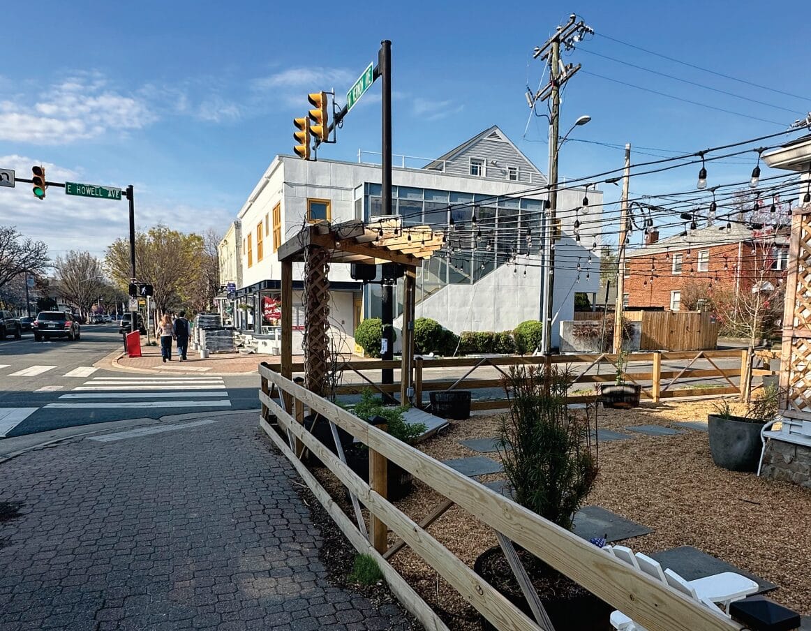 Street view of a small-town street with a crosswalk, storefronts, and string lights overhead on a sunny day.