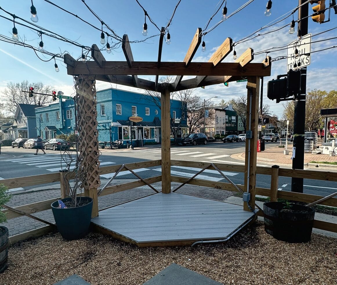 Wooden gazebo deck with string lights over a sidewalk plaza in a small-town street scene.