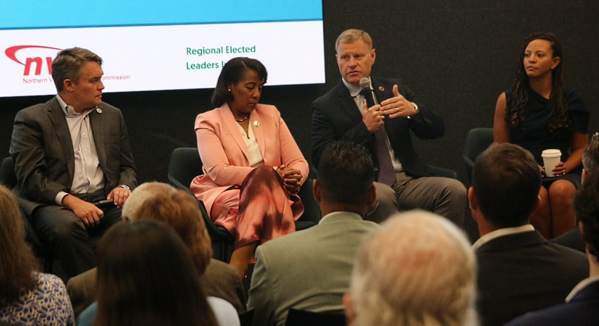 Panel of four adults in business attire seated on stage, one man speaking into a microphone as the audience watches; a slide reads 'Regional Elected Leaders' in the background.